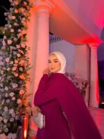 Plum modest cape evening dress with white hijab and silver clutch at wedding venue