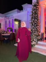 Plum modest cape evening dress with white hijab and silver clutch at wedding venue