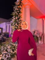 Plum modest cape evening dress with white hijab and silver clutch at wedding venue