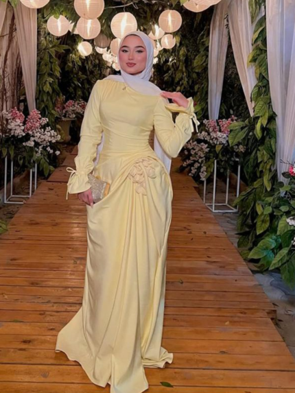 Pastel yellow modest evening dress with long sleeves and draped waist, styled with white hijab in floral wedding venue