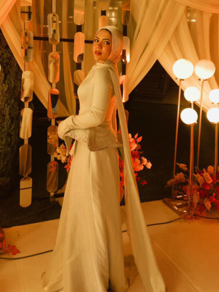 Off-white modest peplum evening dress with long shawl and beige hijab under warm golden party lights