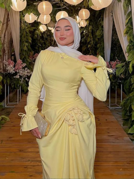 Pastel yellow modest evening dress with long sleeves and draped waist, styled with white hijab in floral wedding venue
